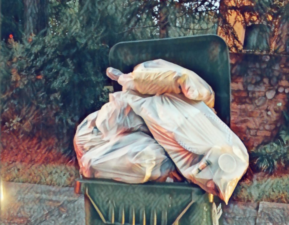 Overstuffed trash bags piled on top of an open green wheelie bin