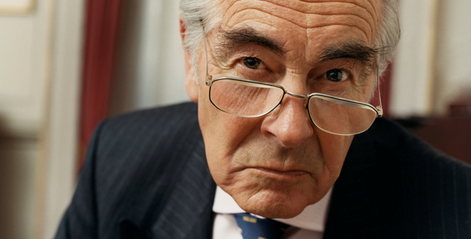An older man in a suit peering sternly over his glasses