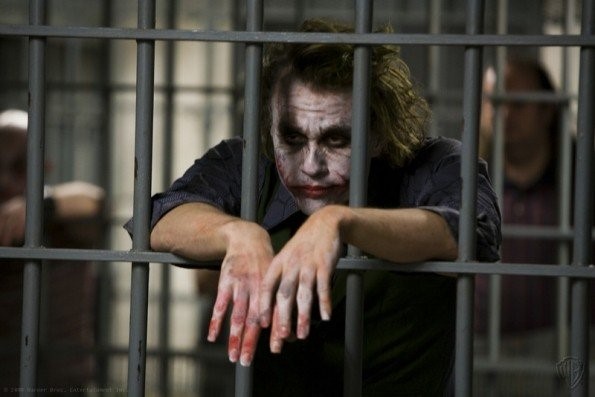 A man in Joker face paint leaning on the bars of a jail cell