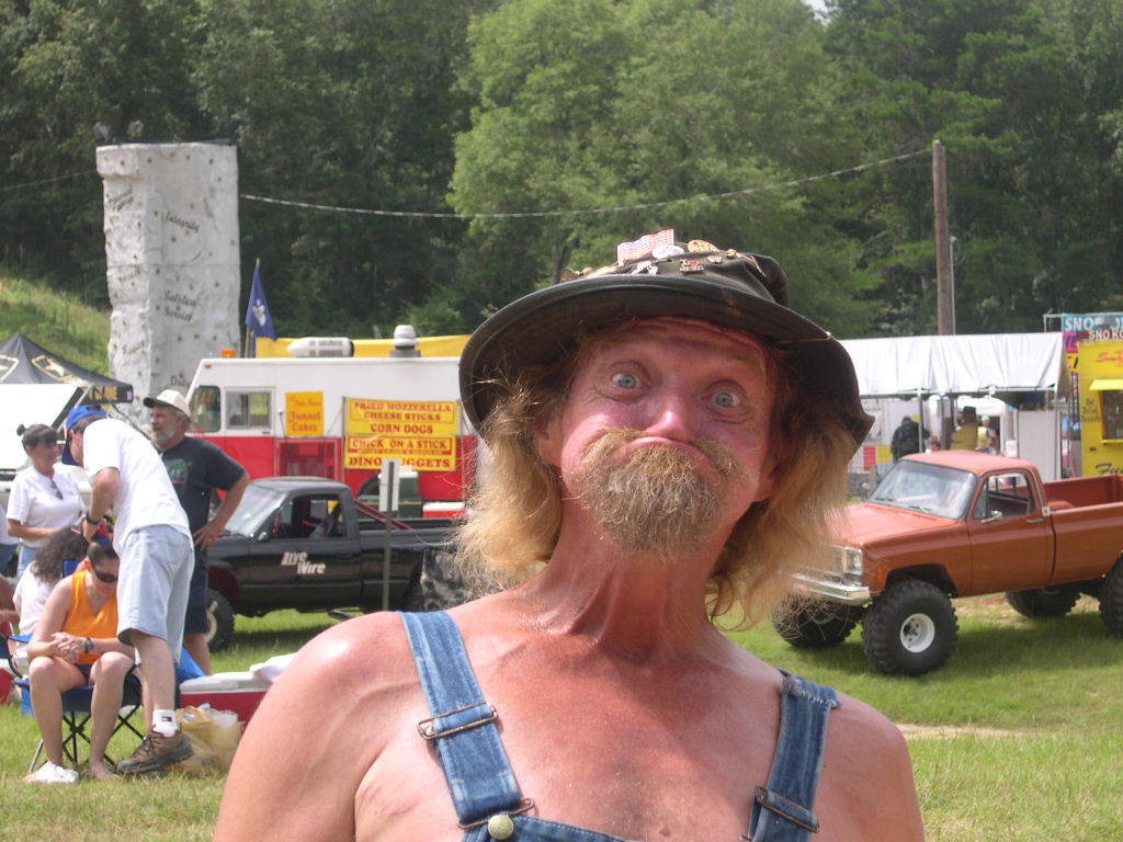 A shirtless man in overalls and a floppy hat with a large mustache, at an outdoor gathering with pickup trucks